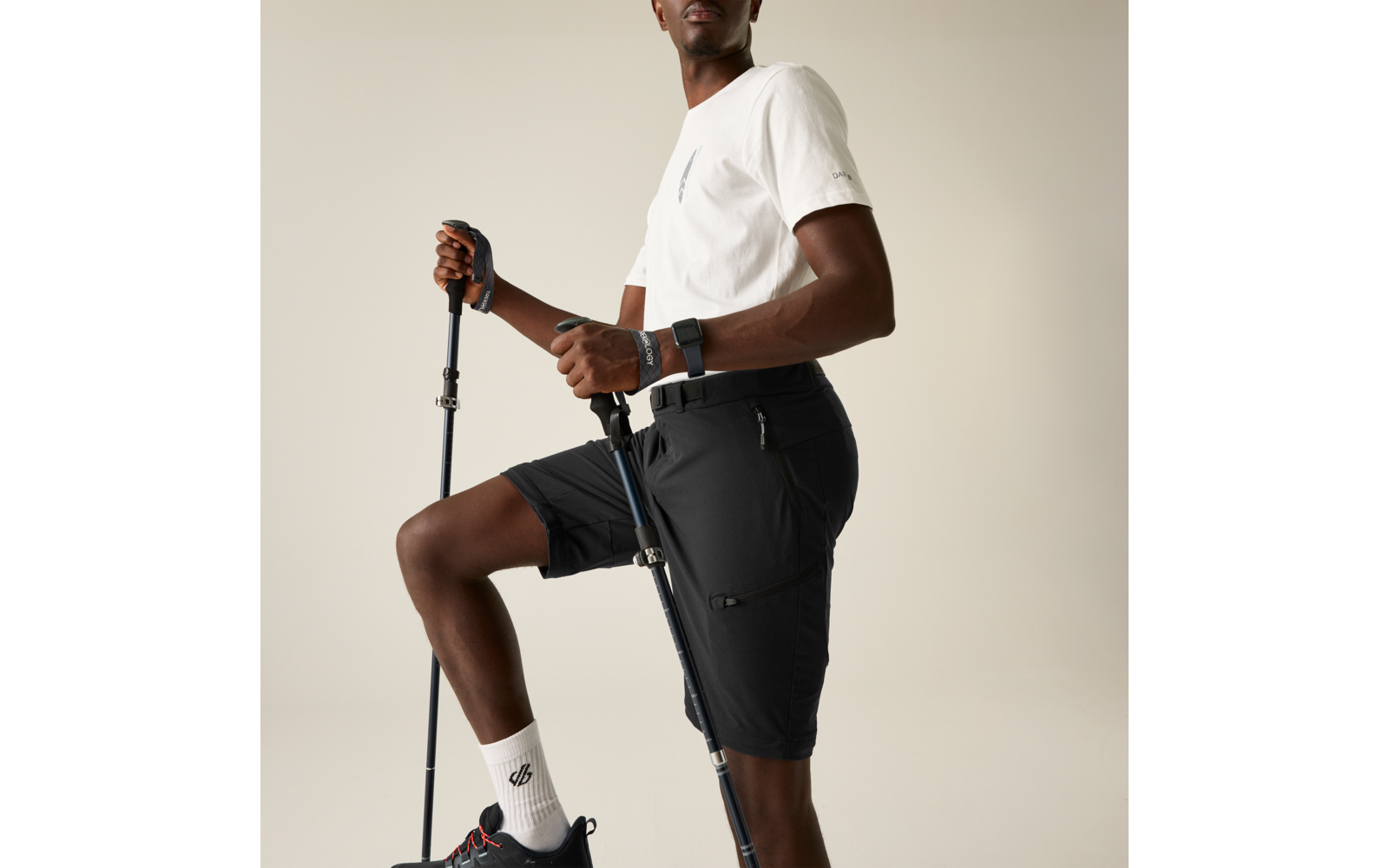 Persona con camiseta blanca y pantalones cortos negros, sostiene dos bastones de senderismo, una rodilla apoyada en una superficie elevada, calcetines blancos, zapatillas negras, reloj inteligente en la muñeca