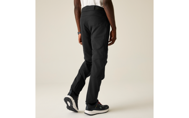 Vista posterior de una persona desde la cintura hacia abajo, pantalones negros con detalles de costura y cremallera en la caña, dobladillo de una camiseta blanca, zapatillas negro y blanco, fondo claro