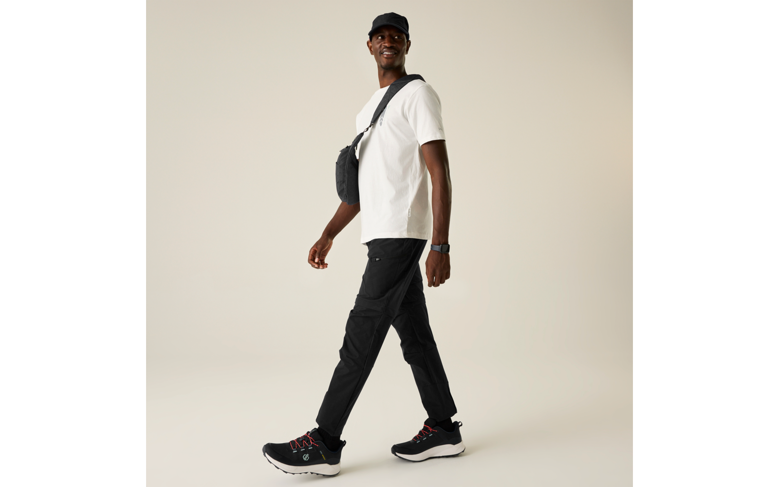 Hombre con camiseta blanca, gorra negra, bolso bandolera negro, pantalones negros y zapatillas negras, camina hacia la derecha sobre fondo claro