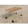 Table rectangulaire avec plateau en bois brun clair et quatre pieds métalliques gris clair munis de plaques de pied rondes noires, posée sur du gravier
