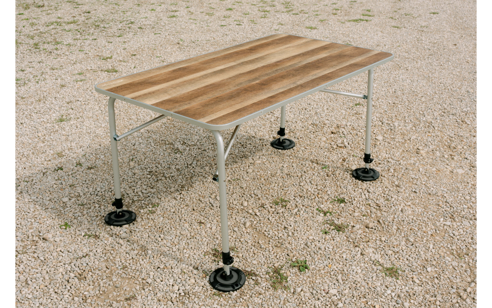 Table rectangulaire avec plateau en bois brun clair et quatre pieds métalliques gris clair munis de plaques de pied rondes noires, posée sur du gravier