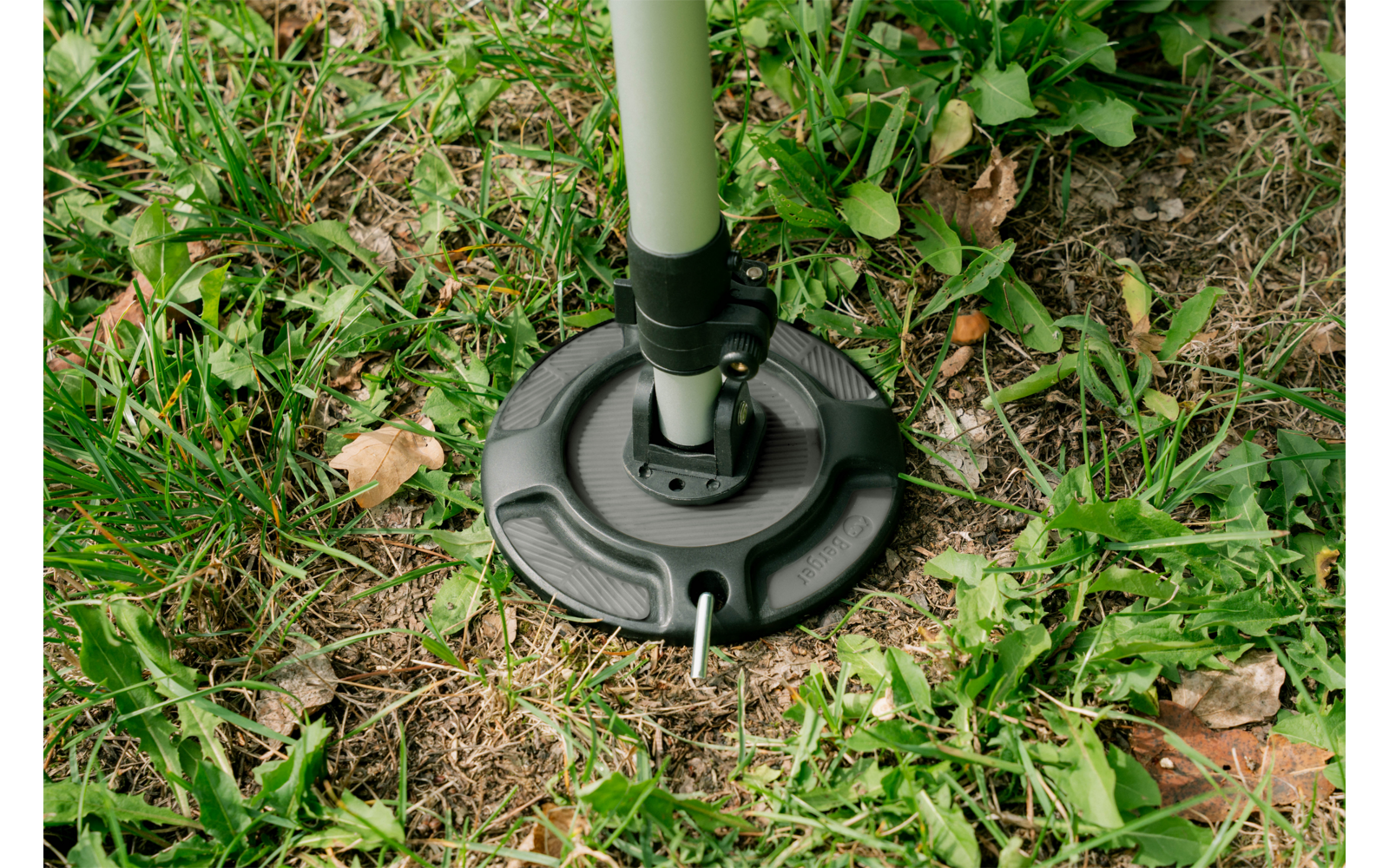 Plaque de base ronde noire avec un poteau gris clair fixé, sur de l'herbe avec de petites feuilles, de la terre et une courte tige métallique à l'avant