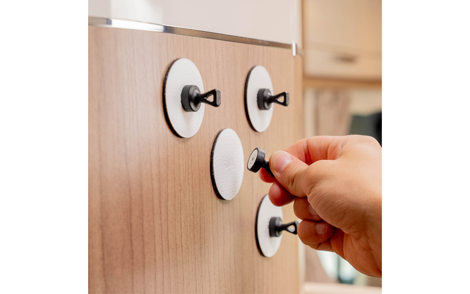 Right hand holds a small black round object against a light brown wood panel with four white round discs and black hooks