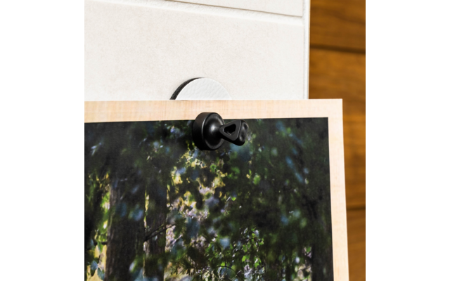 Top edge of a light wood frame with a dark photo area of green tree leaves, attached with a black plastic hook to a white wall; right edge shows brown wood paneling