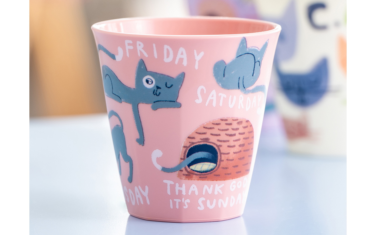Nahaufnahme von drei bunten Bechern auf einer hellen Oberfläche; vorn ein rosa Becher mit blauen Tierzeichnungen und den Worten "FRIDAY", "SATURDAY" und "THANK GOD IT'S SUNDAY"; im Hintergrund unscharf ein violetter und ein weißer Becher