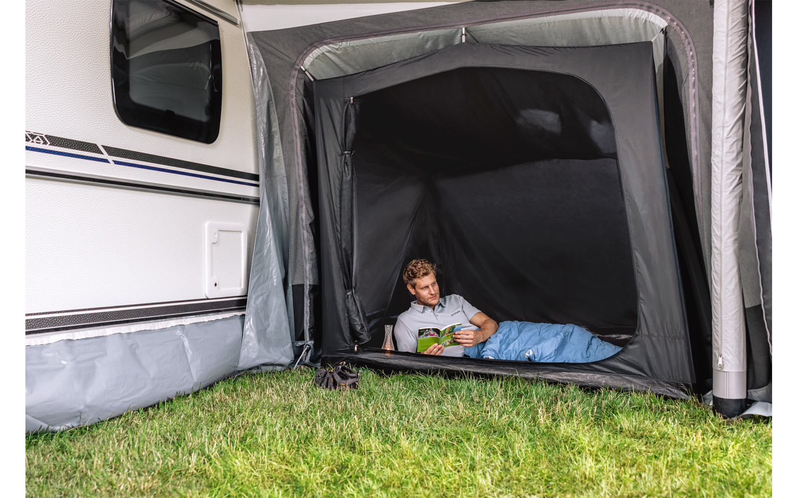 Kind in slaapzak leest boek in voortent van een caravan; gras ervoor