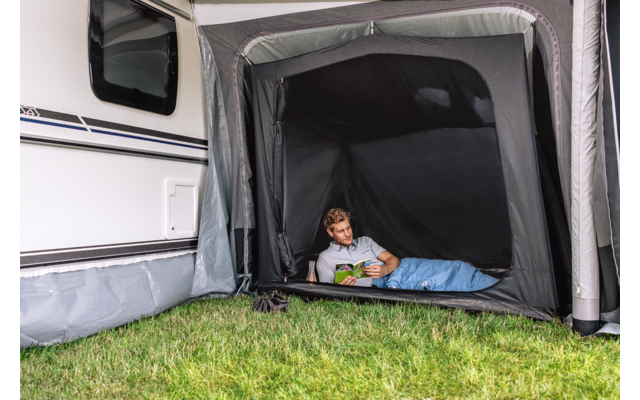 Kind in slaapzak leest boek in voortent van een caravan; gras ervoor