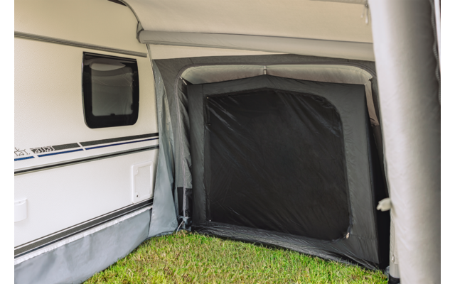 Zijaanzicht van een witte caravan met een aangebouwd grijs voortent, een zwart rechthoekig paneel bij de opening en gras op de grond