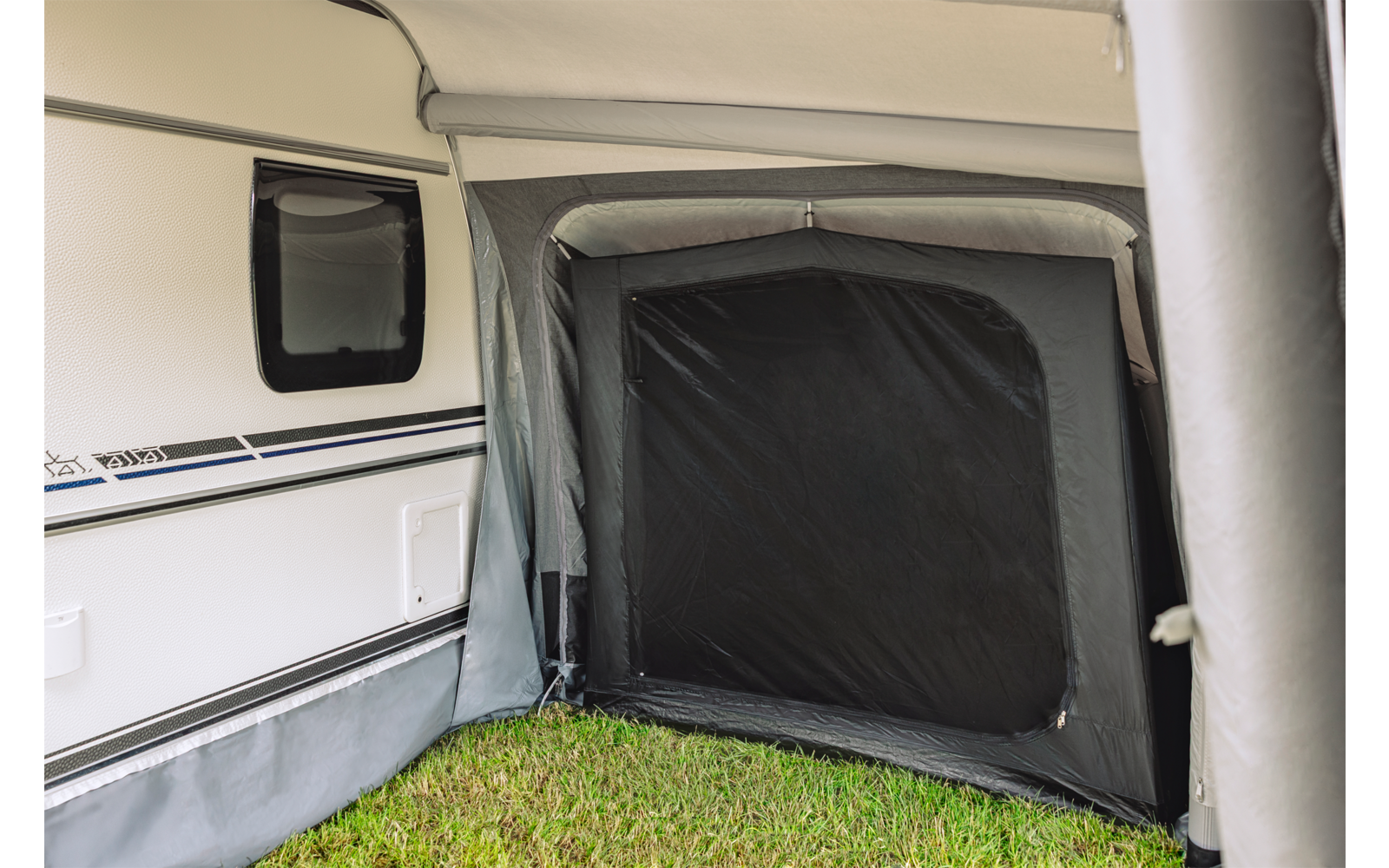Zijaanzicht van een witte caravan met een aangebouwd grijs voortent, een zwart rechthoekig paneel bij de opening en gras op de grond