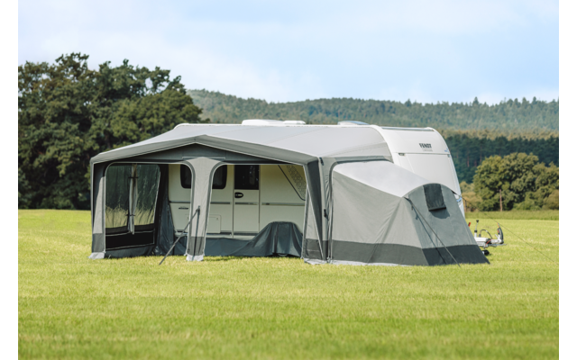 Wit-grijze caravan met aangebouwde grijze voortent op een groen veld, bomen en bewolkte lucht op de achtergrond