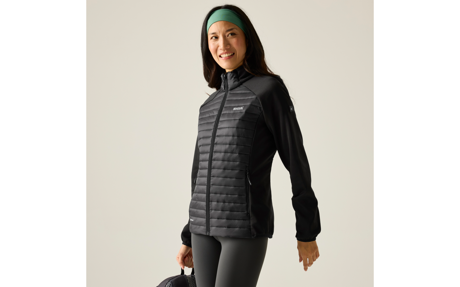 Persoon in driekwartsprofiel draagt een zwarte doorgestikte jas met rits, zwarte legging, een groen haarband en houdt een zwarte tas