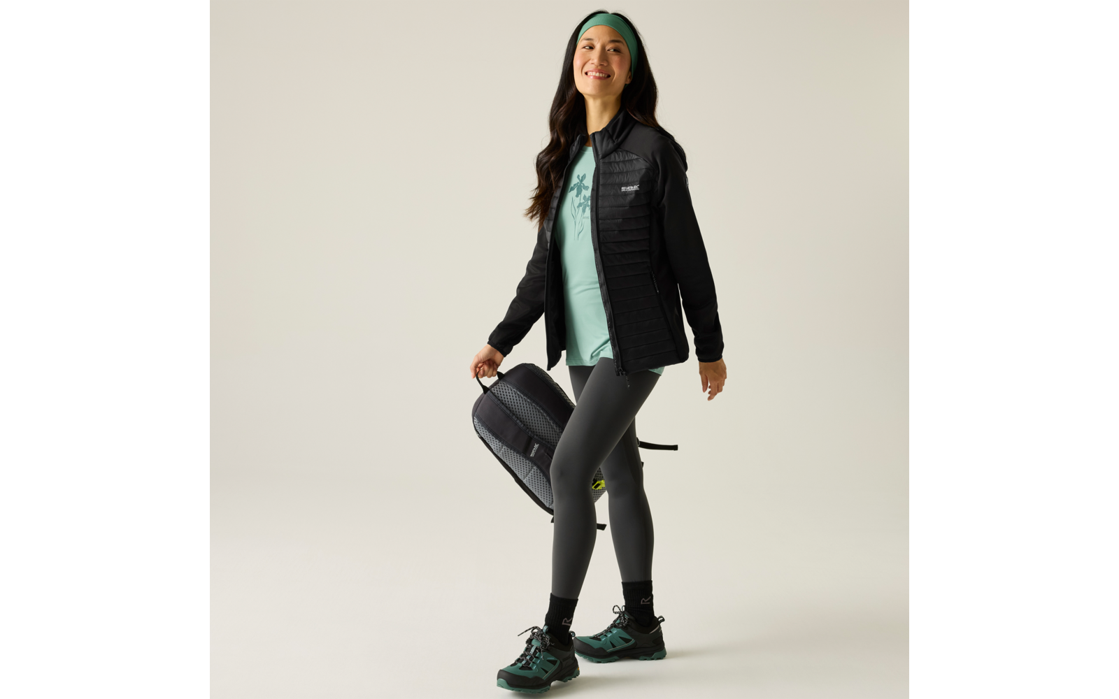 Vrouw staat voor een lichte achtergrond, draagt een gewatteerde zwarte jas, licht turkoois T-shirt, donkere legging, turkooise haarband, houdt een zwarte rugzak in haar rechterhand, draagt turkoois-zwarte wandelschoenen