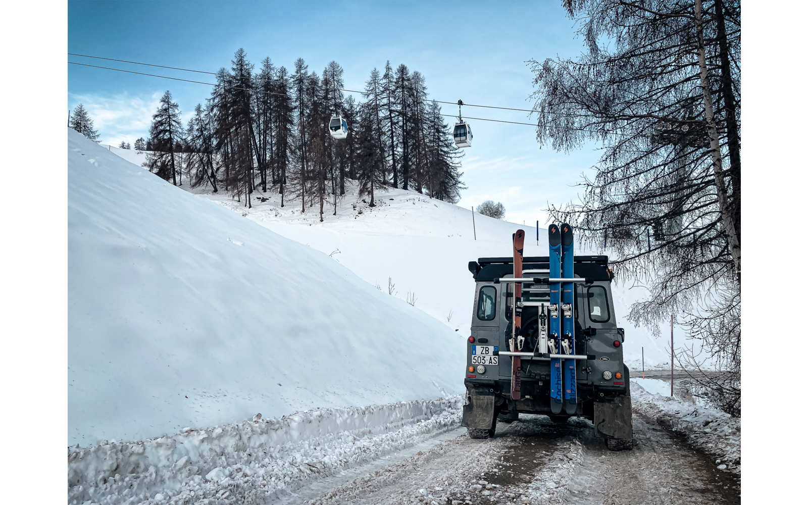 Veicolo con cingoli e sci fissati al retro su una strada innevata, cabine della funivia su un cavo sopra un pendio coperto di neve, conifere e cielo azzurro