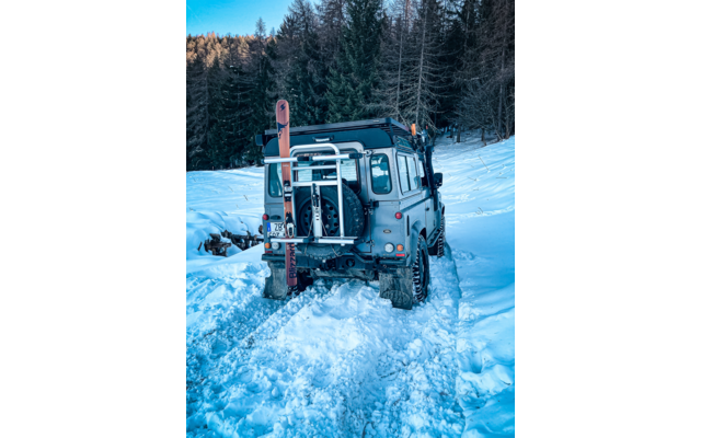 Veicolo fuoristrada visto da dietro su una pista innevata, ruota di scorta e due sci montati sul retro, conifere sullo sfondo