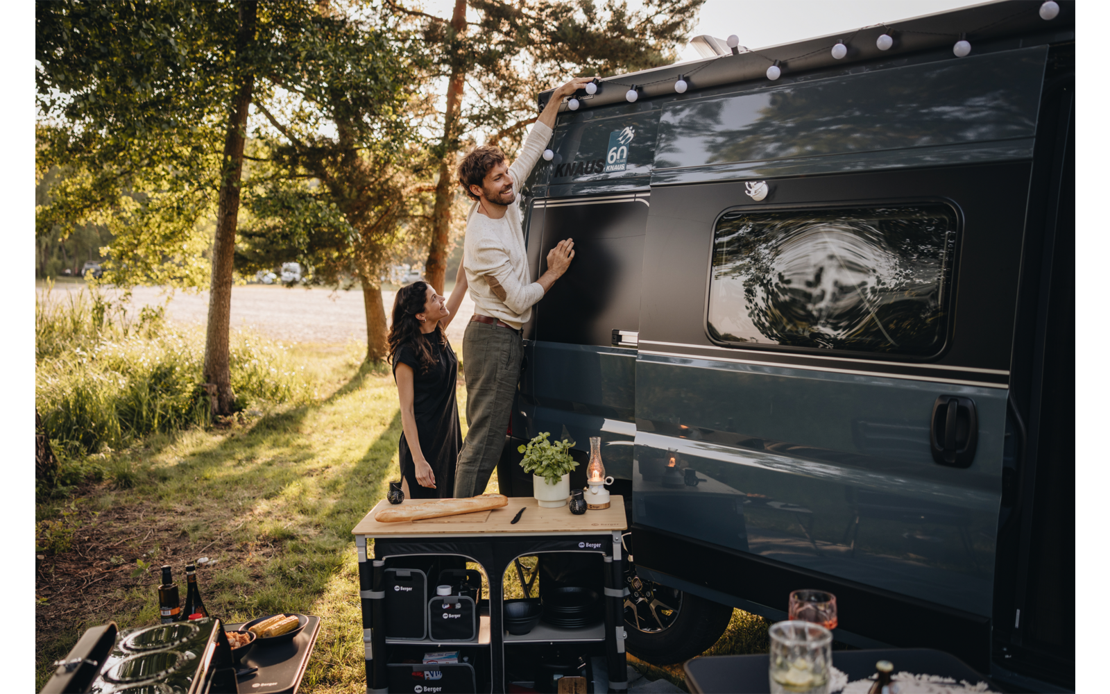 Deux personnes à côté d'un camping-car foncé : l'une est sur une petite marche et tend la main vers l'extérieur du véhicule, l'autre tient la marche ; devant le camping-car une table de camping avec une plante en pot, de la vaisselle et des boissons, en arrière-plan des arbres et un lac