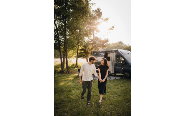 Deux personnes se tiennent la main en marchant sur une pelouse devant un camping-car garé dont la porte est ouverte ; arbres et soleil en arrière-plan