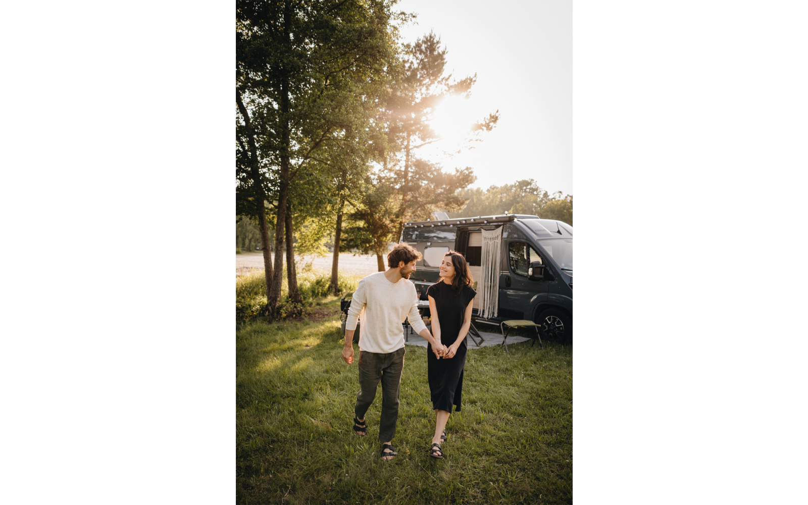 Deux personnes se tiennent la main en marchant sur une pelouse devant un camping-car garé dont la porte est ouverte ; arbres et soleil en arrière-plan