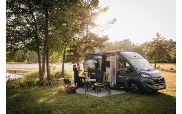 Fourgon aménagé gris à porte latérale ouverte stationné sur une pelouse au bord d’un lac, deux personnes à côté, table pliante et chaises de camping, arbres et lumière du soleil en arrière-plan