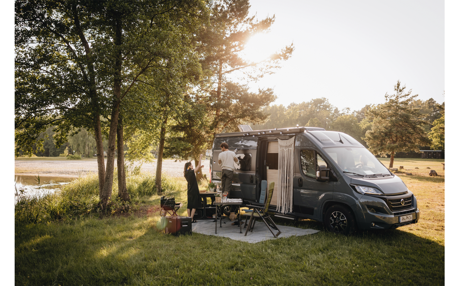 Fourgon aménagé gris à porte latérale ouverte stationné sur une pelouse au bord d’un lac, deux personnes à côté, table pliante et chaises de camping, arbres et lumière du soleil en arrière-plan
