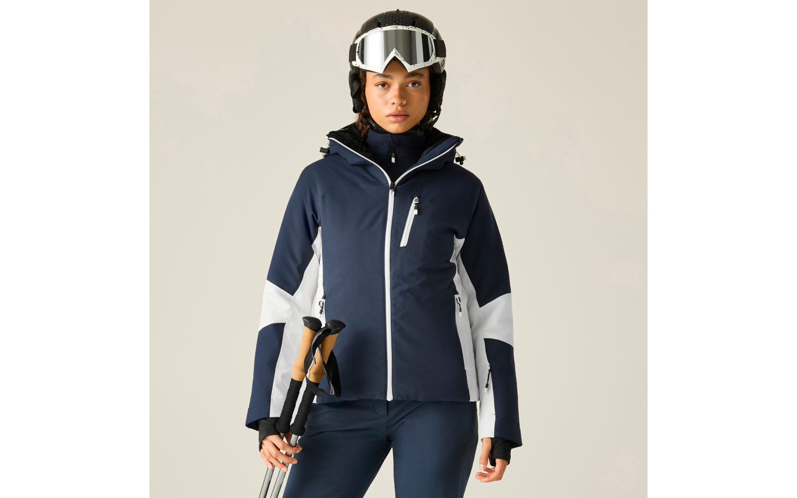 Person in dunkelblauer Skijacke mit weißen Einsätzen, trägt Helm und Skibrille, hält Skistöcke, steht frontal vor hellem Hintergrund