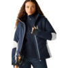 Person mit lockigen Haaren, dunkelblaue Jacke mit weißen Einsätzen, schwarzer Pullover mit Reißverschluss, dunkle Hose, hält Skistöcke