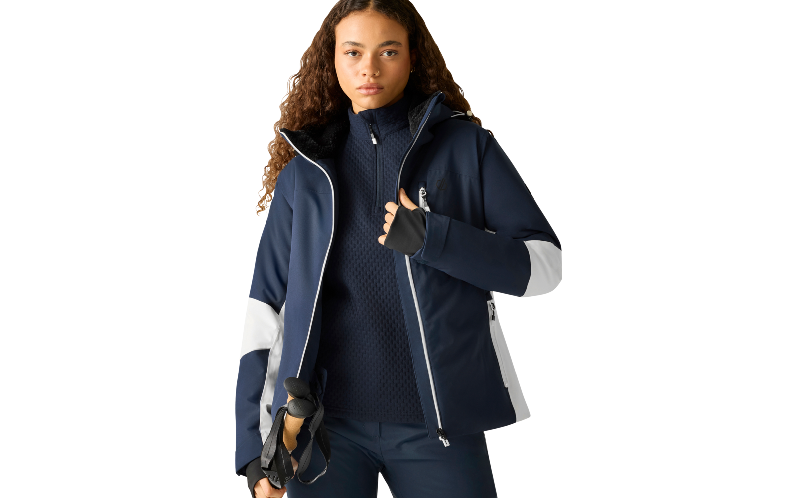 Person mit lockigen Haaren, dunkelblaue Jacke mit weißen Einsätzen, schwarzer Pullover mit Reißverschluss, dunkle Hose, hält Skistöcke