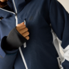 Nahaufnahme: dunkelblaue Jacke mit weißen Reißverschlüssen und seitlichem weißen Einsatz; Hand mit Daumenloch-Bündchen zieht an der Reißverschluss der Brusttasche