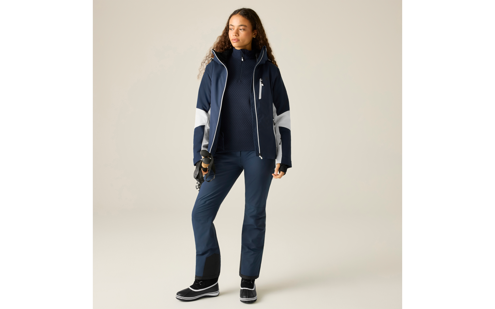 Person mit langen lockigen Haaren trägt eine dunkelblaue Jacke mit Kapuze und weißen Einsätzen, dunkelblaue Hose und schwarze Stiefel, hält Skibrille und Handschuhe in der rechten Hand, steht vor hellem Hintergrund und blickt zur Seite