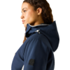Seitliches Porträt einer Person mit lockigem braunem Haar, die eine dunkelblaue Kapuzenjacke mit Kordelzug und goldene Ohrstecker trägt