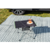 Pequeña mesa de camping plegable con superficie negra y borde verde; sobre ella un vaso transparente con líquido de color naranja sobre un paño blanco con pequeñas flores blancas; la mesa está sobre una manta de picnic gris con patrón de rombos blancos; al fondo césped y parte de un coche con rueda