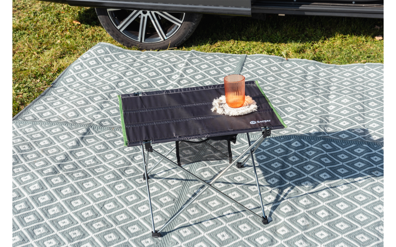 Pequeña mesa de camping plegable con superficie negra y borde verde; sobre ella un vaso transparente con líquido de color naranja sobre un paño blanco con pequeñas flores blancas; la mesa está sobre una manta de picnic gris con patrón de rombos blancos; al fondo césped y parte de un coche con rueda