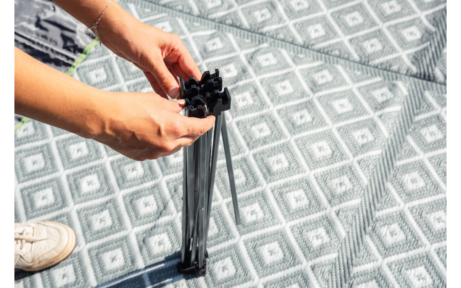 Dos manos sostienen un conector de plástico negro y varillas metálicas plegadas sobre una alfombra gris con patrón de rombos; en la esquina inferior izquierda se ve una zapatilla blanca