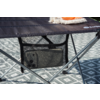 Mesa plegable oscura con bolsillo de malla que contiene un teléfono; patas cruzadas de metal; alfombra gris con patrón debajo; esquina de una manta beige y un objeto naranja sobre la mesa