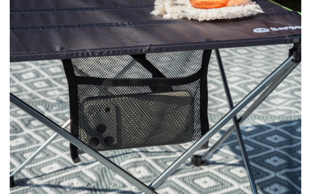 Mesa plegable oscura con bolsillo de malla que contiene un teléfono; patas cruzadas de metal; alfombra gris con patrón debajo; esquina de una manta beige y un objeto naranja sobre la mesa