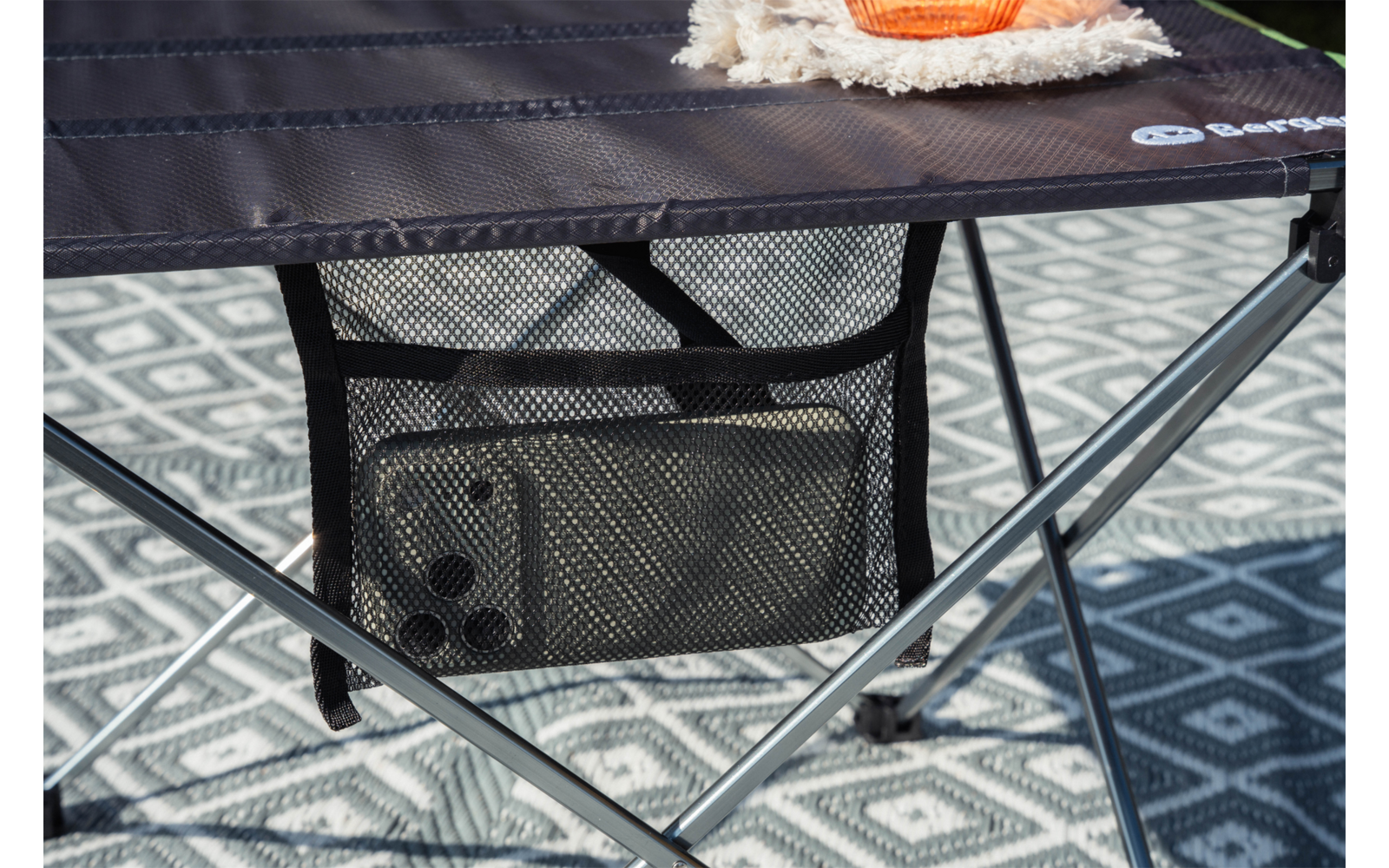 Mesa plegable oscura con bolsillo de malla que contiene un teléfono; patas cruzadas de metal; alfombra gris con patrón debajo; esquina de una manta beige y un objeto naranja sobre la mesa