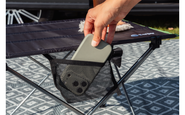 Mano derecha coloca un objeto gris rectangular de bordes redondeados con tres aberturas circulares en un bolsillo de malla negro en una superficie de tela negra con soportes metálicos; debajo se ve una alfombra de patrón gris y blanco