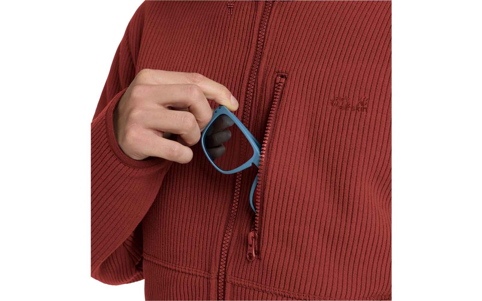 Hand hält blaue Sonnenbrille in geöffneter Brusttasche eines roten, gerippten Reißverschluss-Pullovers