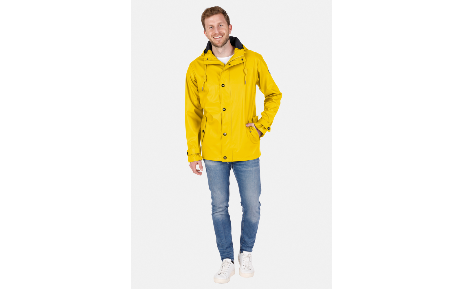 Person steht in gelber Regenjacke, blauen Jeans und weißen Sneakern auf hellem Hintergrund, lächelnd zur Kamera