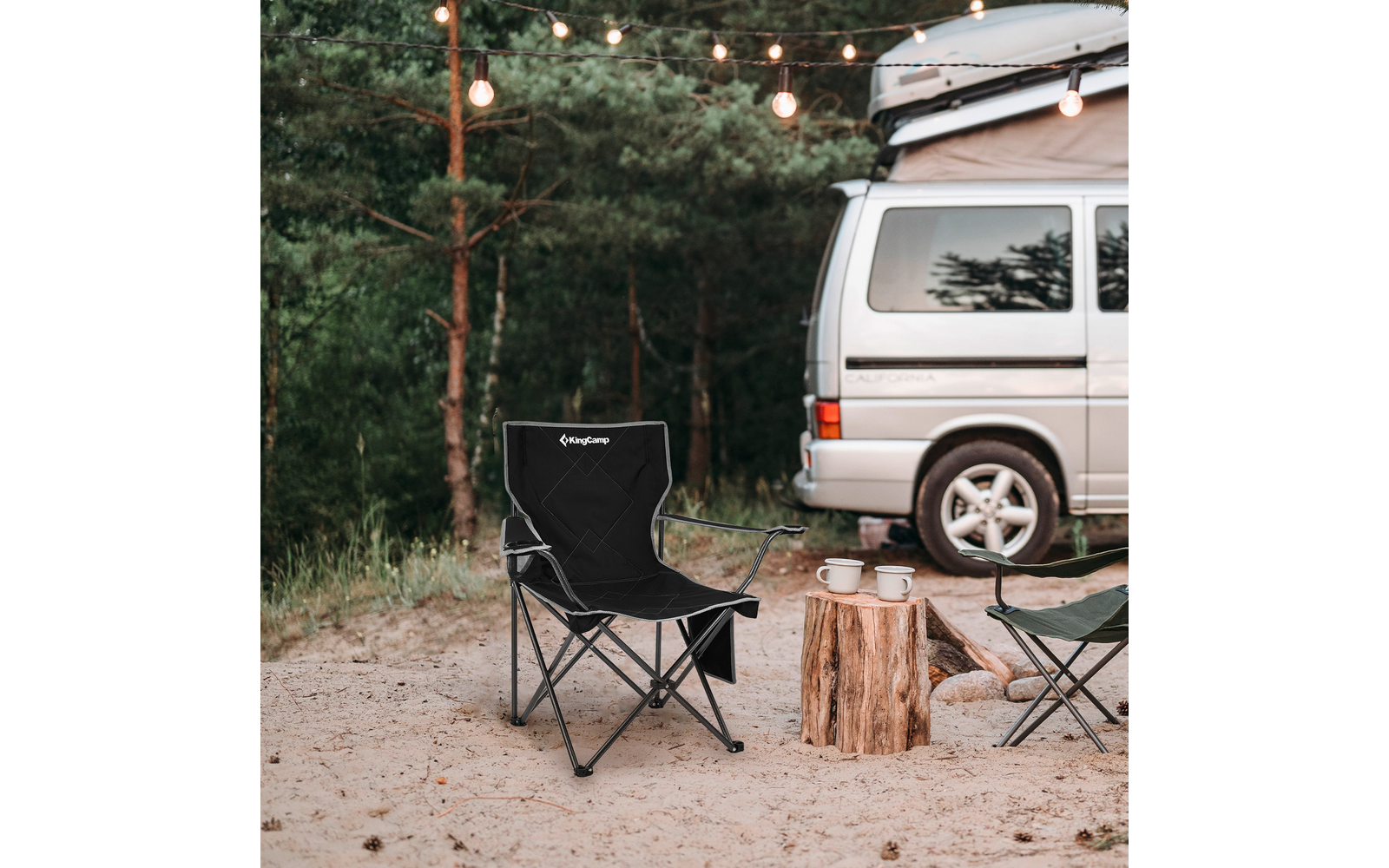Campingplatz mit weißem Van, schwarzem Klappstuhl, Baumstümpfen und Lichterkette in Wäldern