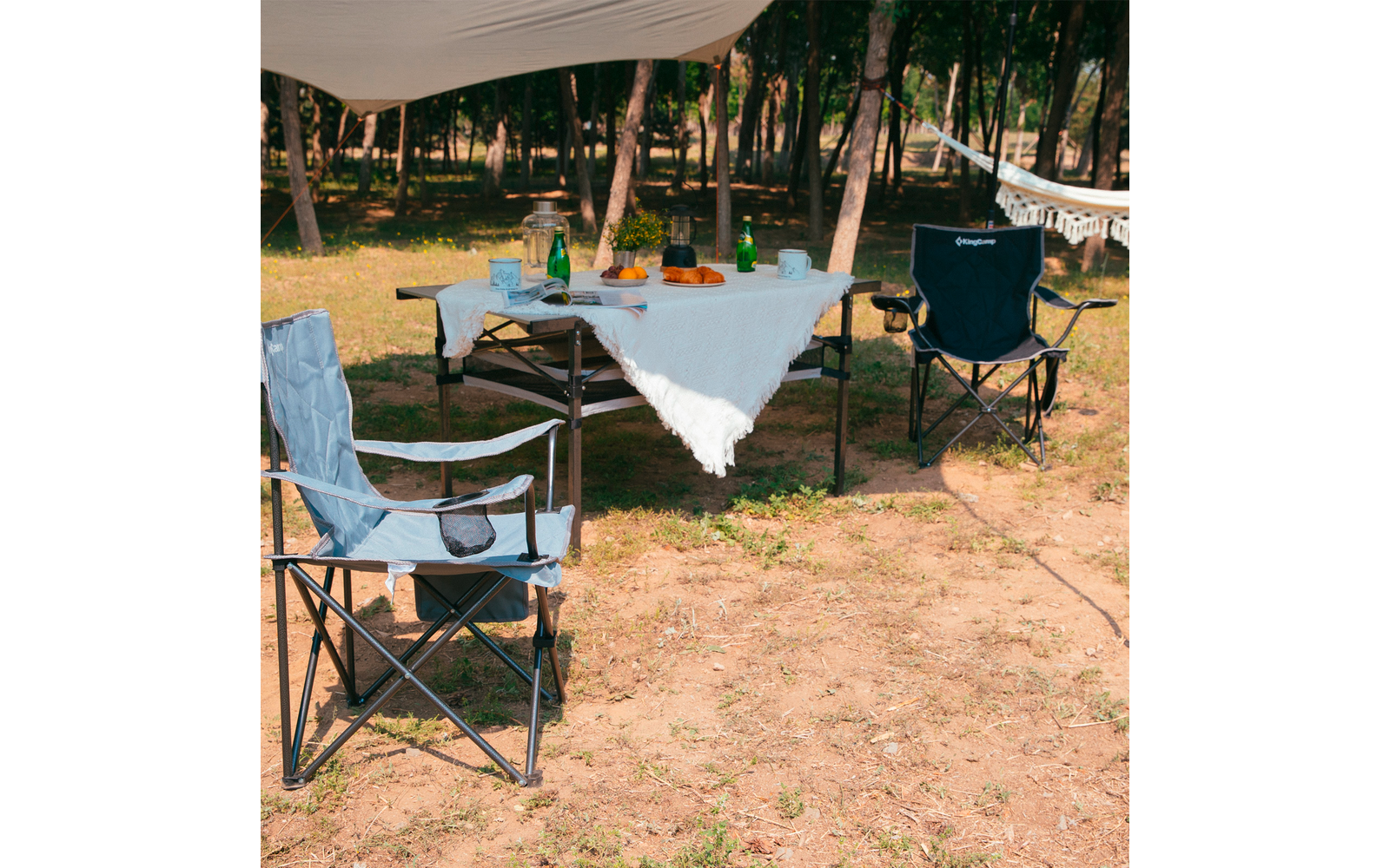 Zwei Klappstühle und ein mit einer weißen Tischdecke bedeckter Klapptisch im Freien auf sandigem Boden, im Hintergrund Bäume