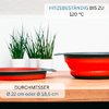 Eine zusammengeklappte und eine aufgeklappte rote Schale mit grauem Rand auf einer Holzfläche; zwei weiße Pflanztöpfe mit grünen Gräsern im Hintergrund; sichtbarer Text: 'HITZEBESTÄNDIG BIS ZU 120 °C' und 'DURCHMESSER Ø 22 cm oder Ø 18,5 cm'