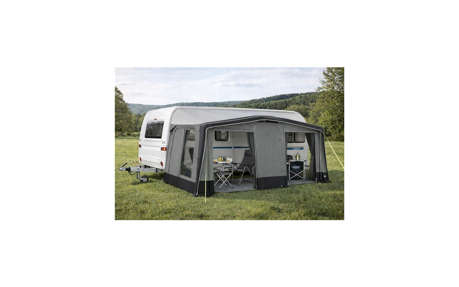Gran toldo de camping gris con varias ventanas rectangulares, franja inferior oscura, cortinas en las ventanas, techo curvo, tensores y estacas; adhesivo redondo rojo abajo a la derecha con texto blanco