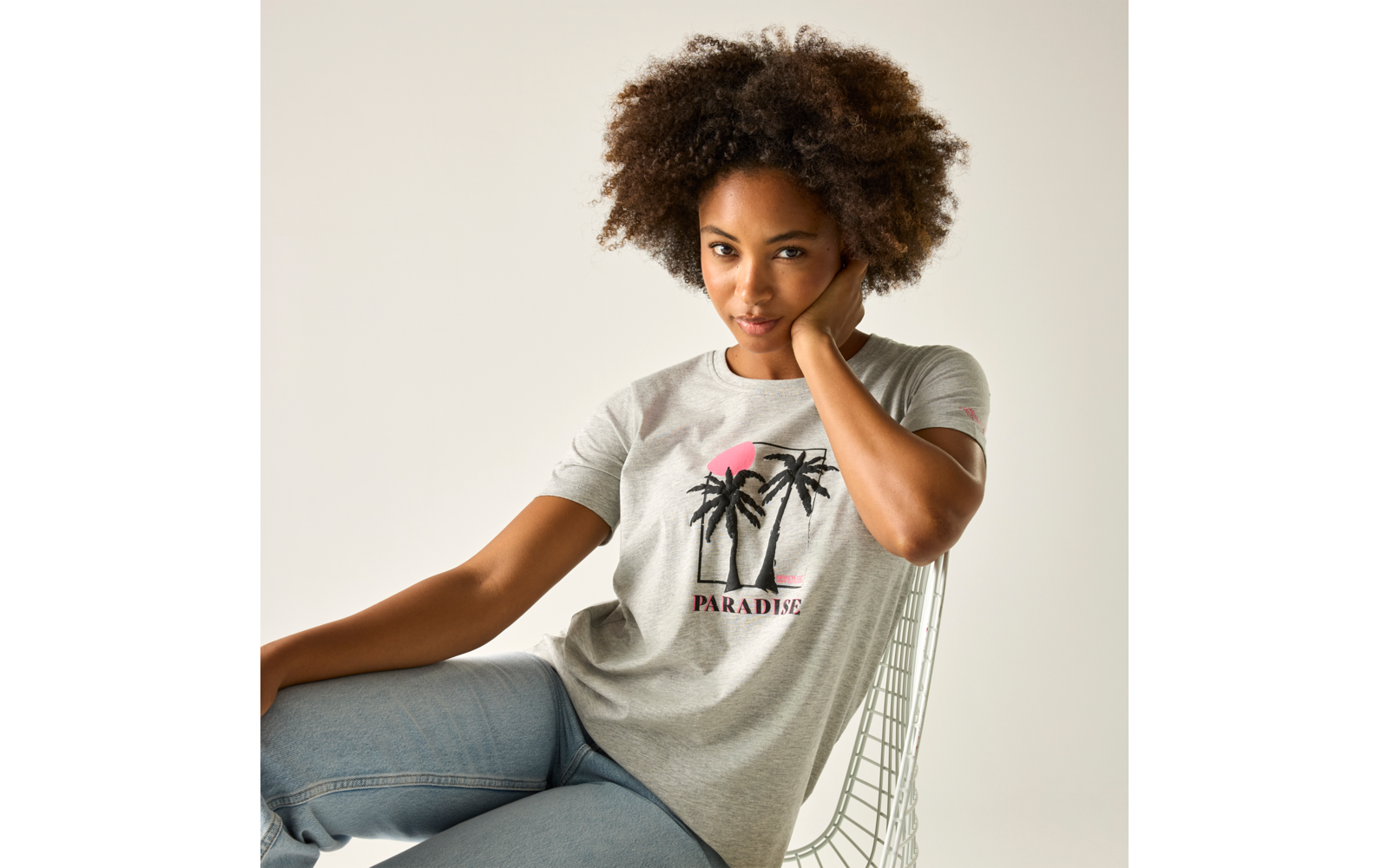 Person sitzt auf einem Drahtstuhl, trägt graues T-Shirt mit Palmenmotiv und Jeans, rechte Hand am Hals, heller Hintergrund