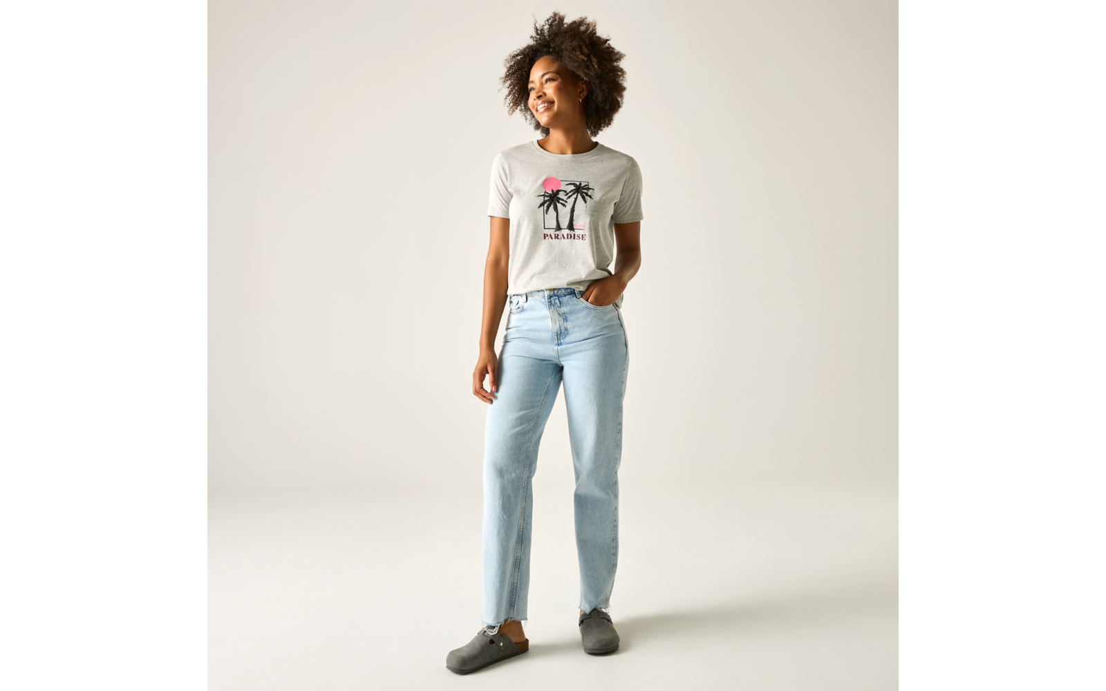 Stehende Person mit lockigen Haaren, lächelnd, trägt weißes T‑Shirt mit Palmenprint, hellblaue Jeans und graue Schuhe vor hellem Hintergrund