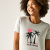 Lächelnde Person mit lockigem Haar trägt hellgraues T-Shirt mit Palmenmotiv, rosafarbenem Kreis und Schriftzug „PARADISE“