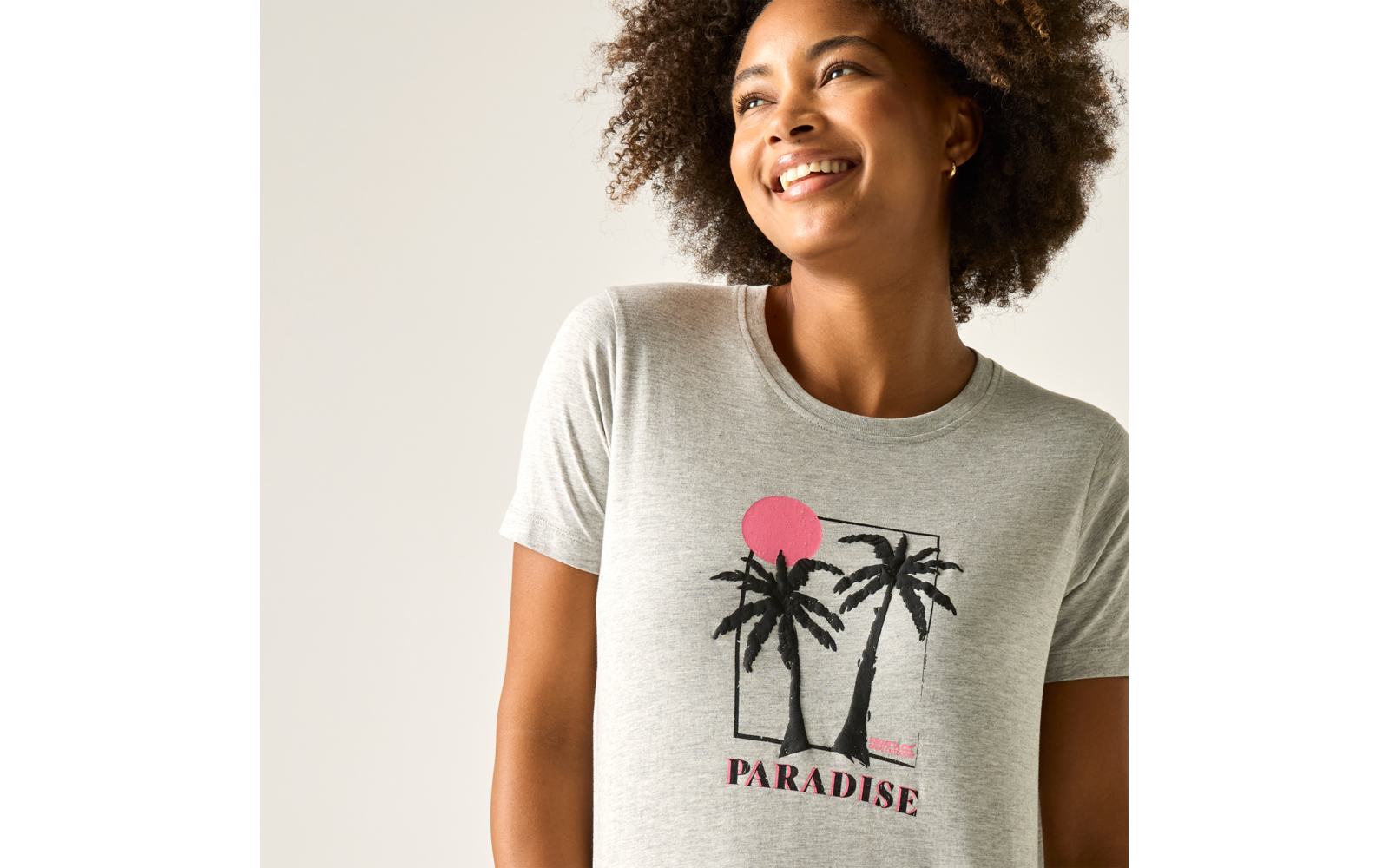 Lächelnde Person mit lockigem Haar trägt hellgraues T-Shirt mit Palmenmotiv, rosafarbenem Kreis und Schriftzug „PARADISE“