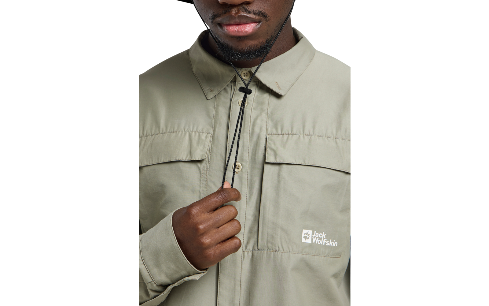 Oberer Torso und untere Gesichtspartie einer Person in heller khakifarbener Jacke mit zwei Brusttaschen und 'Jack Wolfskin'-Logo; eine Hand hält einen schwarzen Kordelzug am Kragen