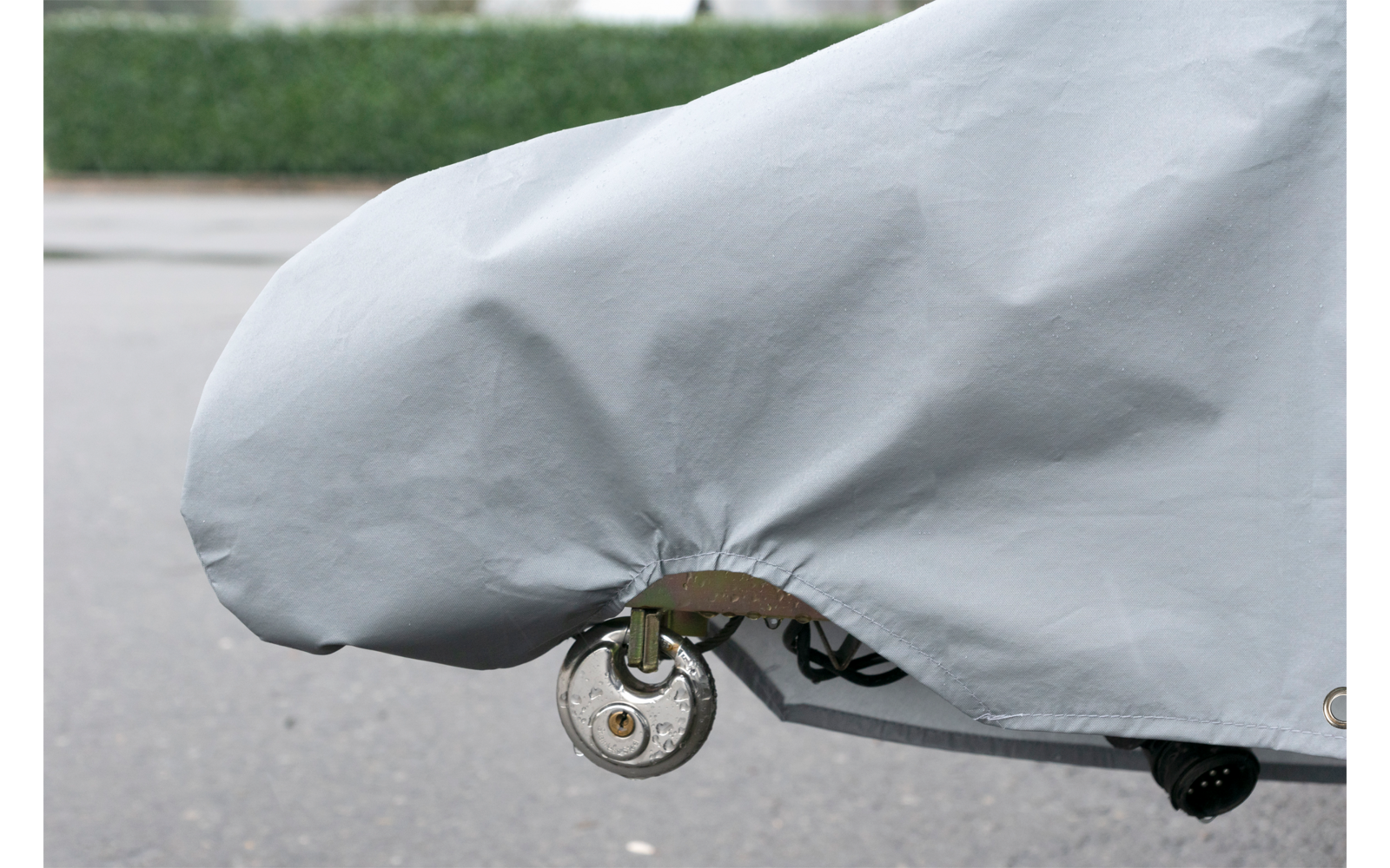Graue Abdeckplane über einem Gegenstand, unten ein freiliegendes Rad und Metallteile, Asphaltboden und Hecke im Hintergrund