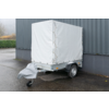 Kleiner Anhänger mit weißer Plane, sichtbares Rad und abgedeckte Deichsel, neben einer grauen gewellten Wand auf nassem Asphalt