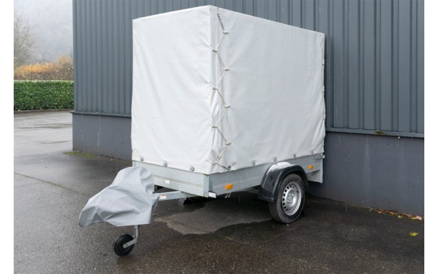 Kleiner Anhänger mit weißer Plane, sichtbares Rad und abgedeckte Deichsel, neben einer grauen gewellten Wand auf nassem Asphalt